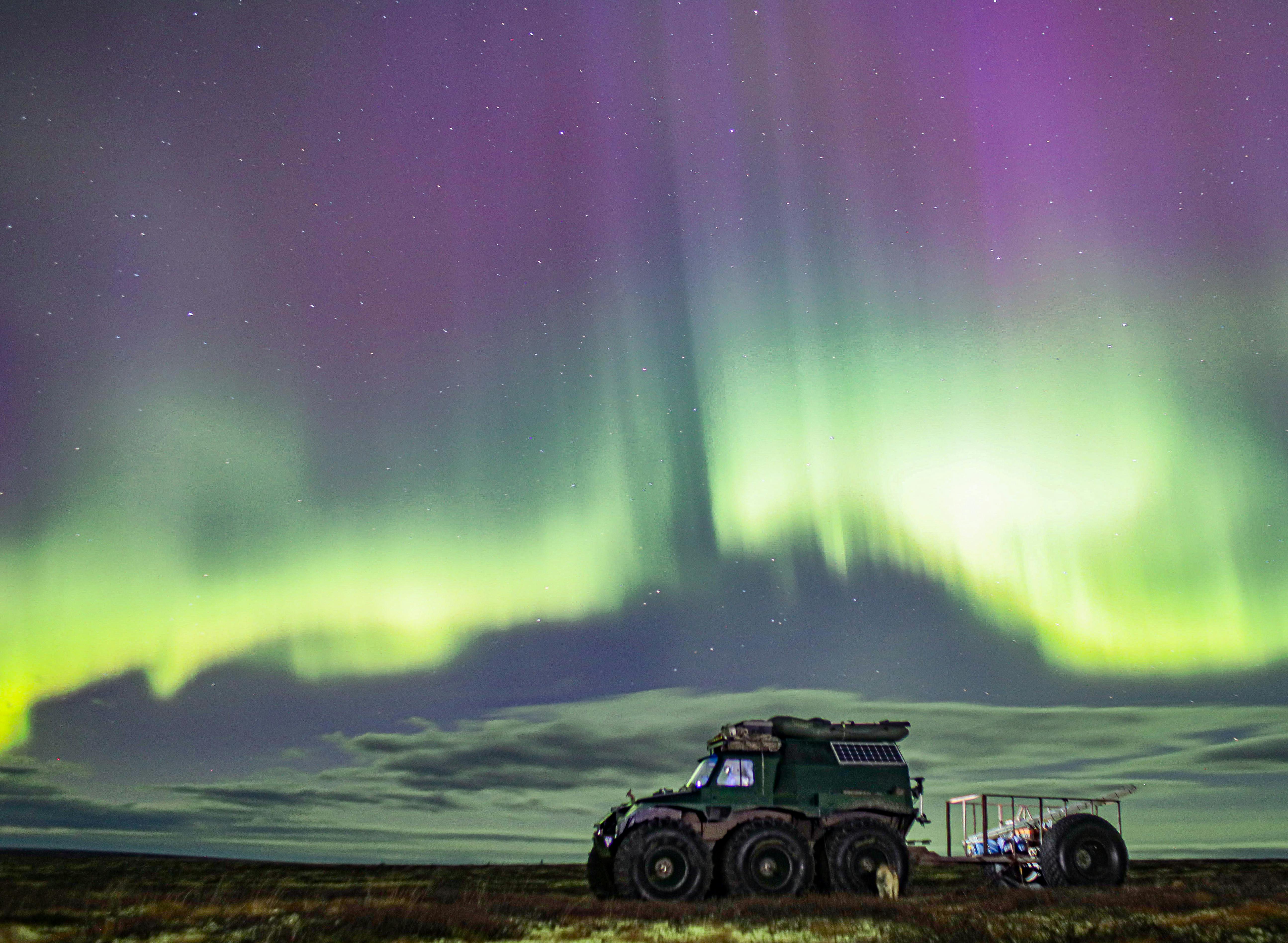 A rugged expedition vehicle sits beneath a vast Arctic sky as ribbons of green and violet light ripple overhead